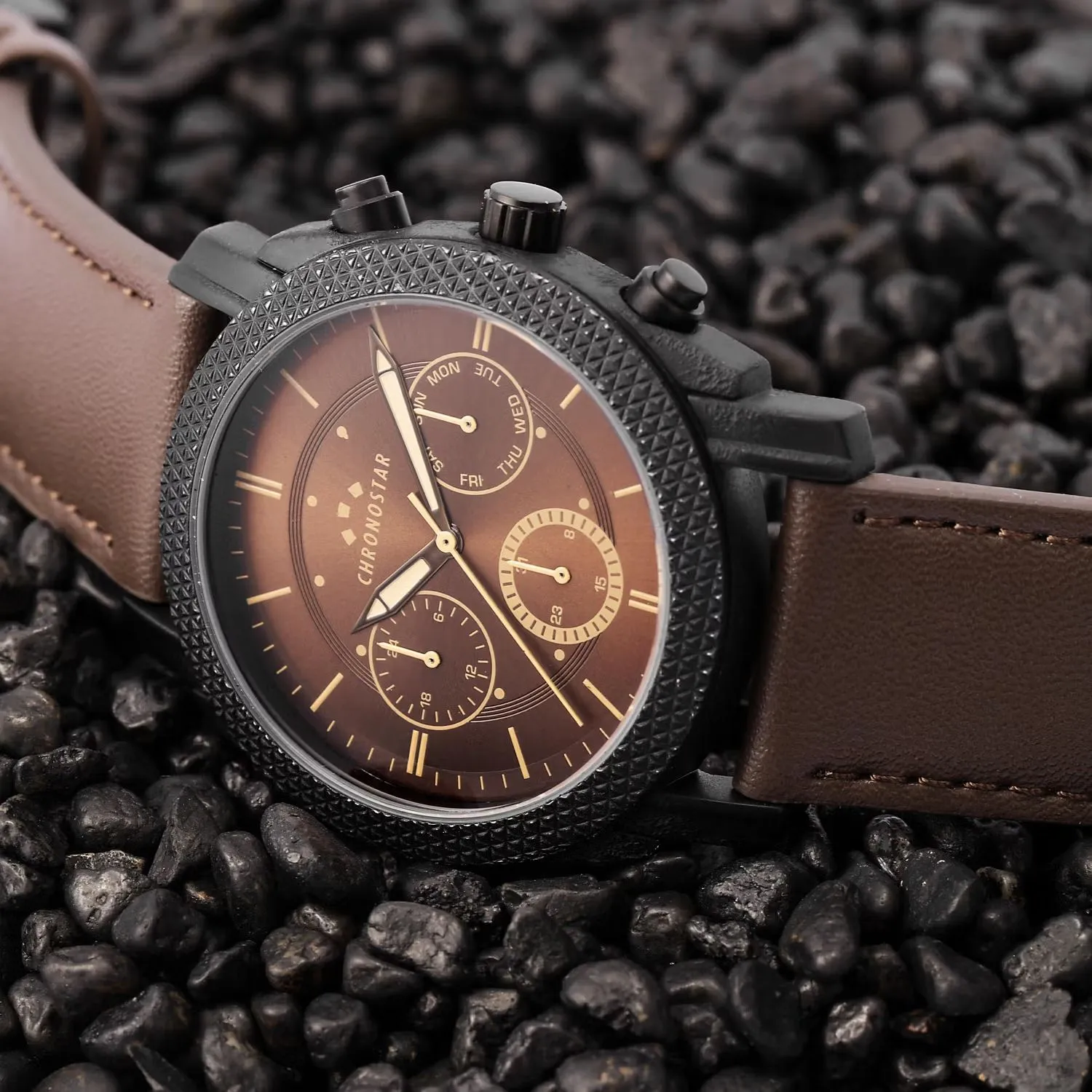 Orologio nero con quadrante marrone e cinturino in pelle, appoggiato su ciottoli scuri.