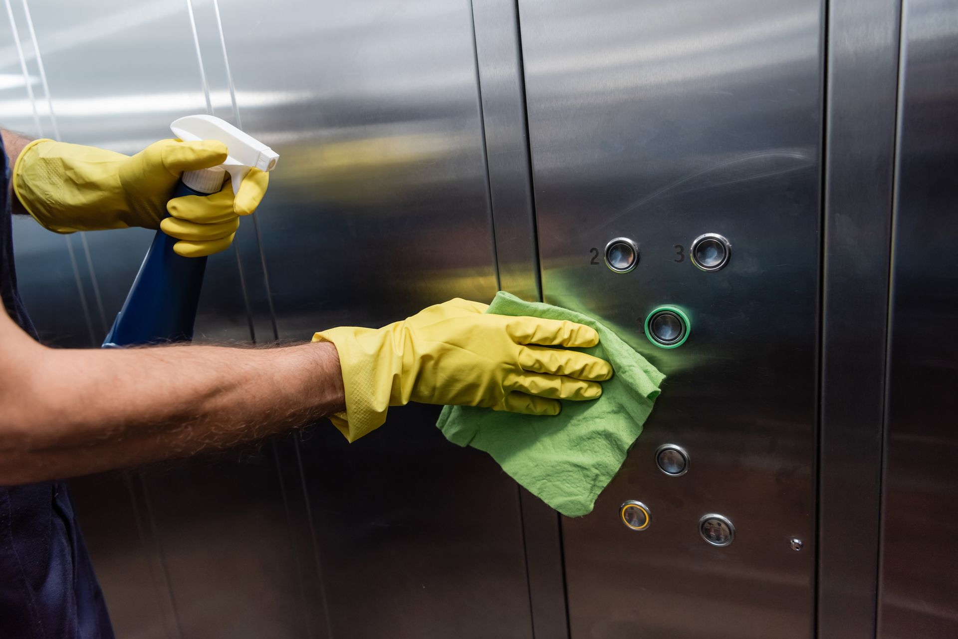 A man wearing yellow gloves is cleaning an elevator with a cloth and spray bottle.
