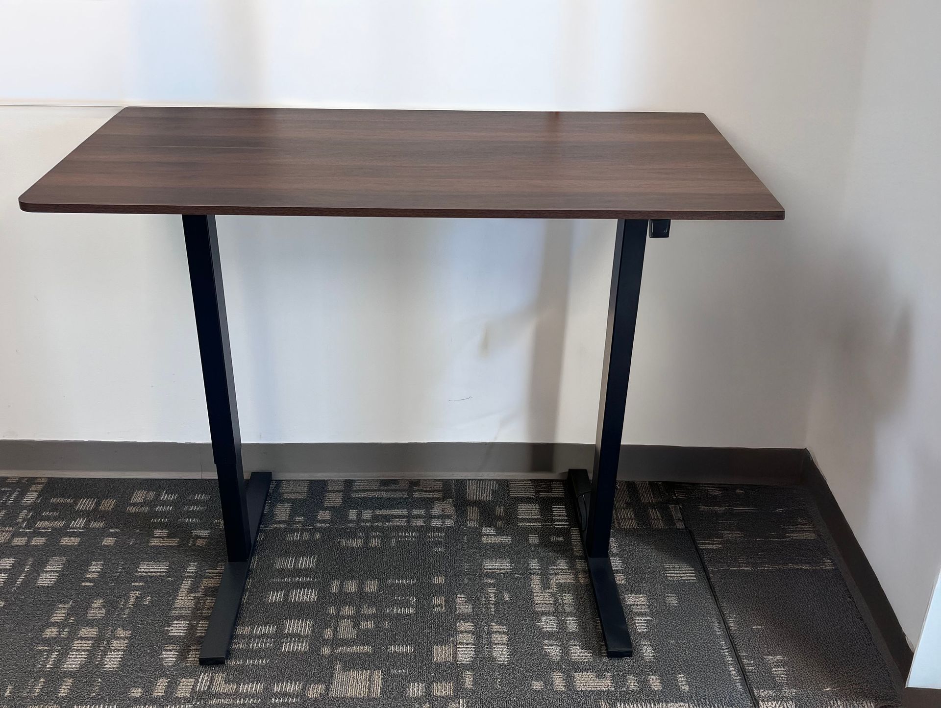 Brown rectangular desk with black legs on patterned carpet against white wall.
