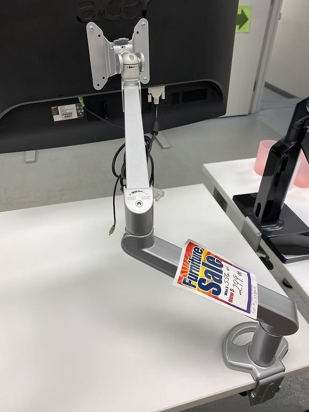 Silver monitor arm clamped to a white desk, supporting a black computer monitor. Sale sign attached.