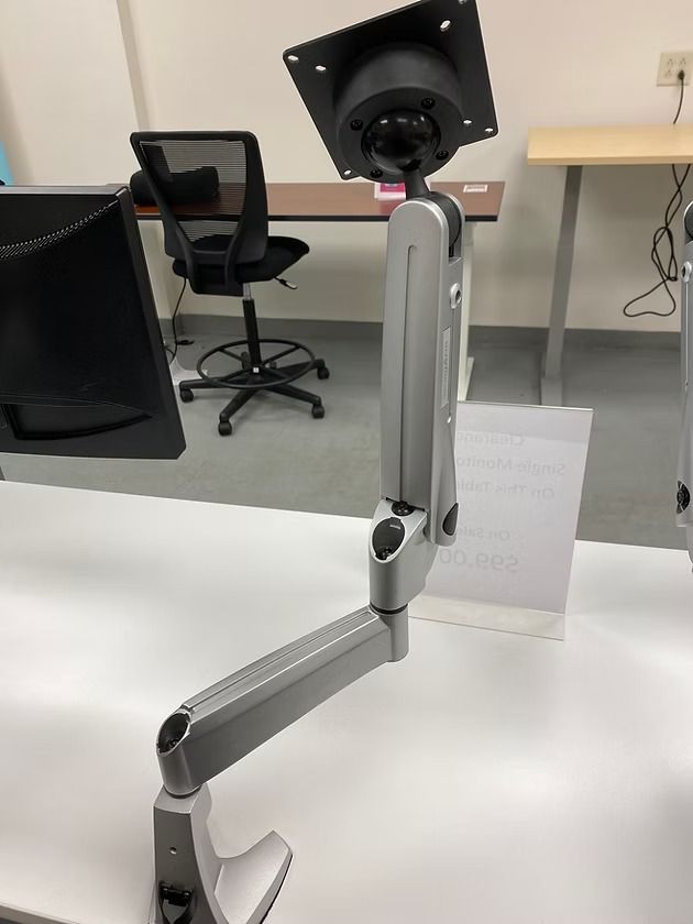 Silver monitor arm extending from a white desk, supporting a screen.