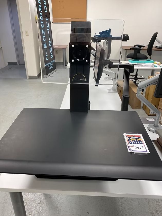 Black adjustable desk with monitor mount and clear shield in a showroom.