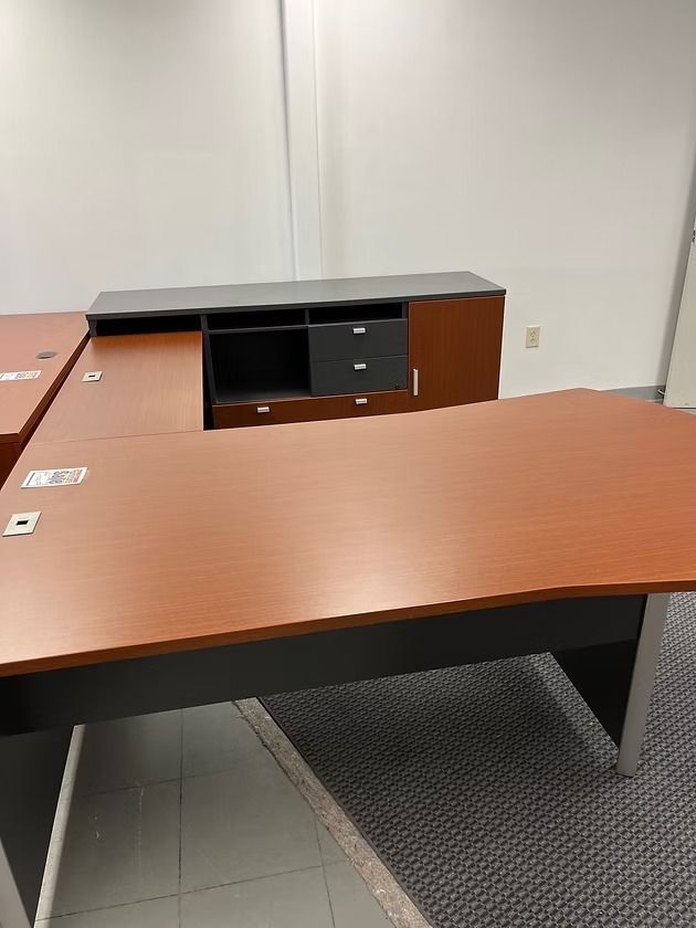 L-shaped office desk, brown top, black trim, with attached cabinet. Grey carpet and white wall background.