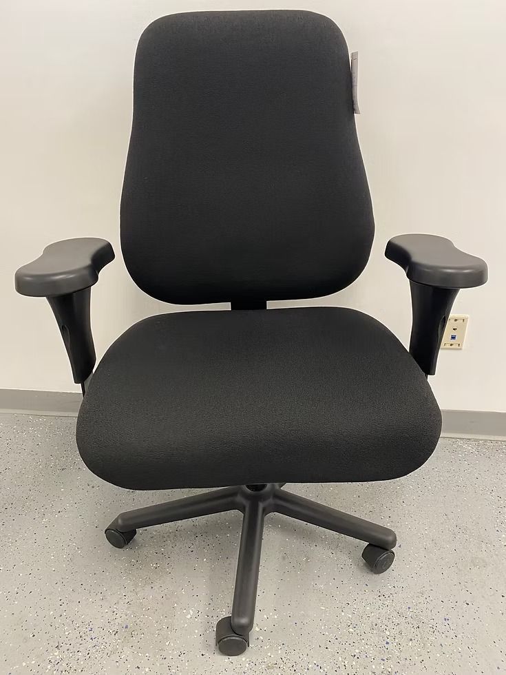 Black office chair with adjustable armrests, against a light gray wall and floor.