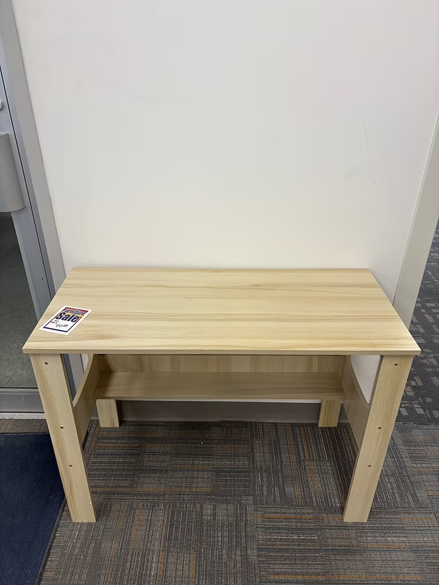 Wooden desk with a shelf, set against a white wall and carpeted floor.