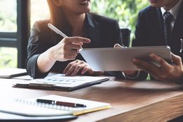 Two people in business attire reviewing documents and a tablet at a table.