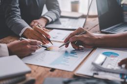 People in business suits analyze charts and graphs at a wooden table.