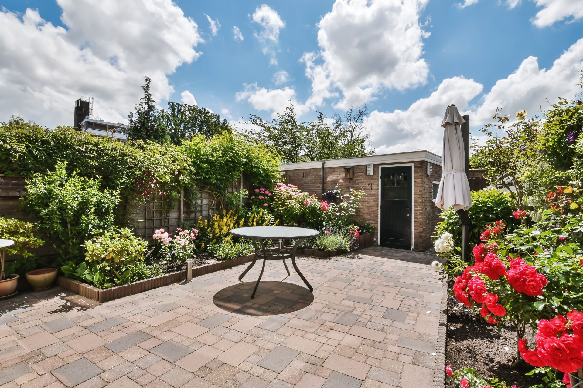 A paved patio with a round table, lush greenery, and blooming red roses under a sunny, cloudy sky.