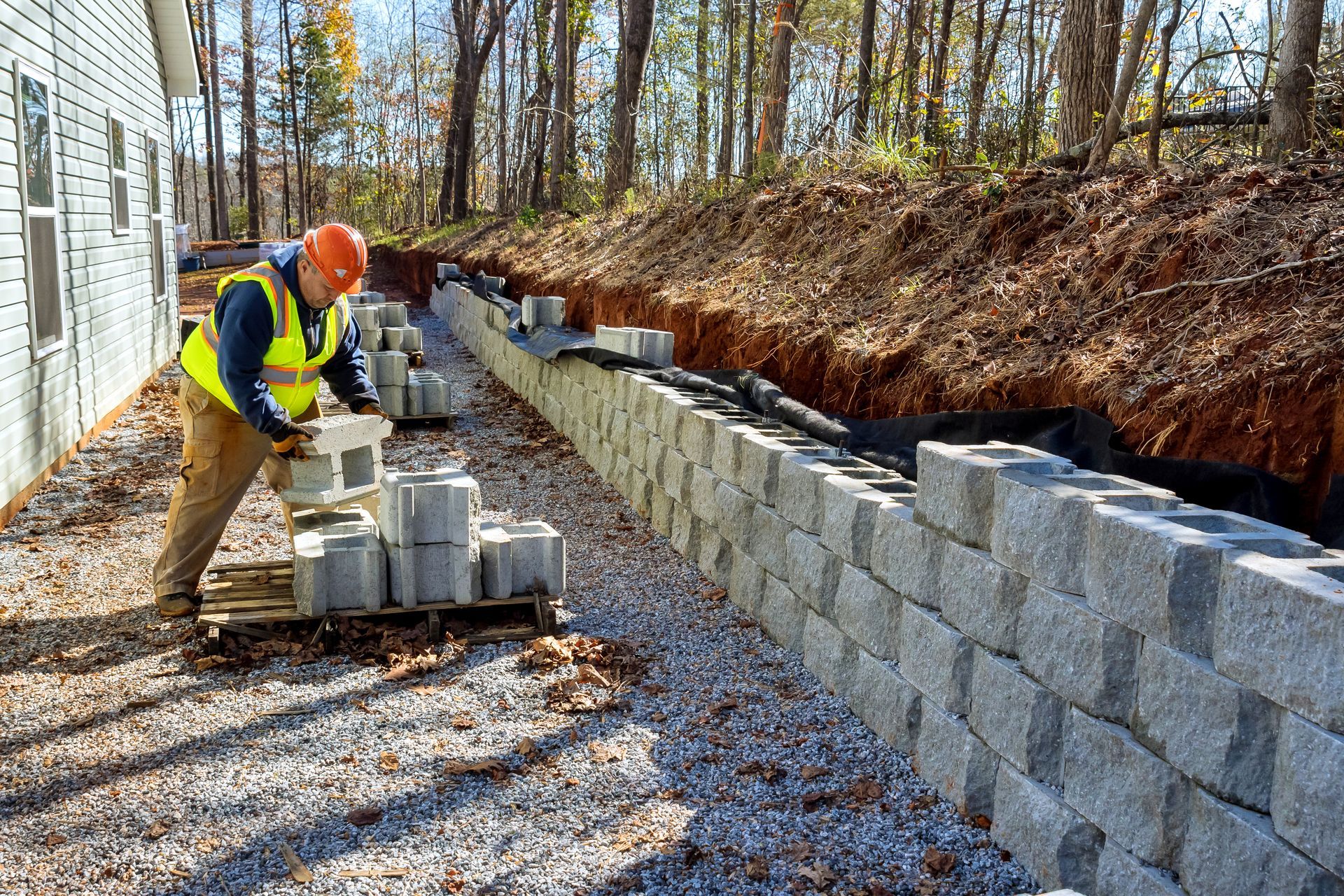 Construction worker building a retaining wall with concrete blocks near a building and hillside.