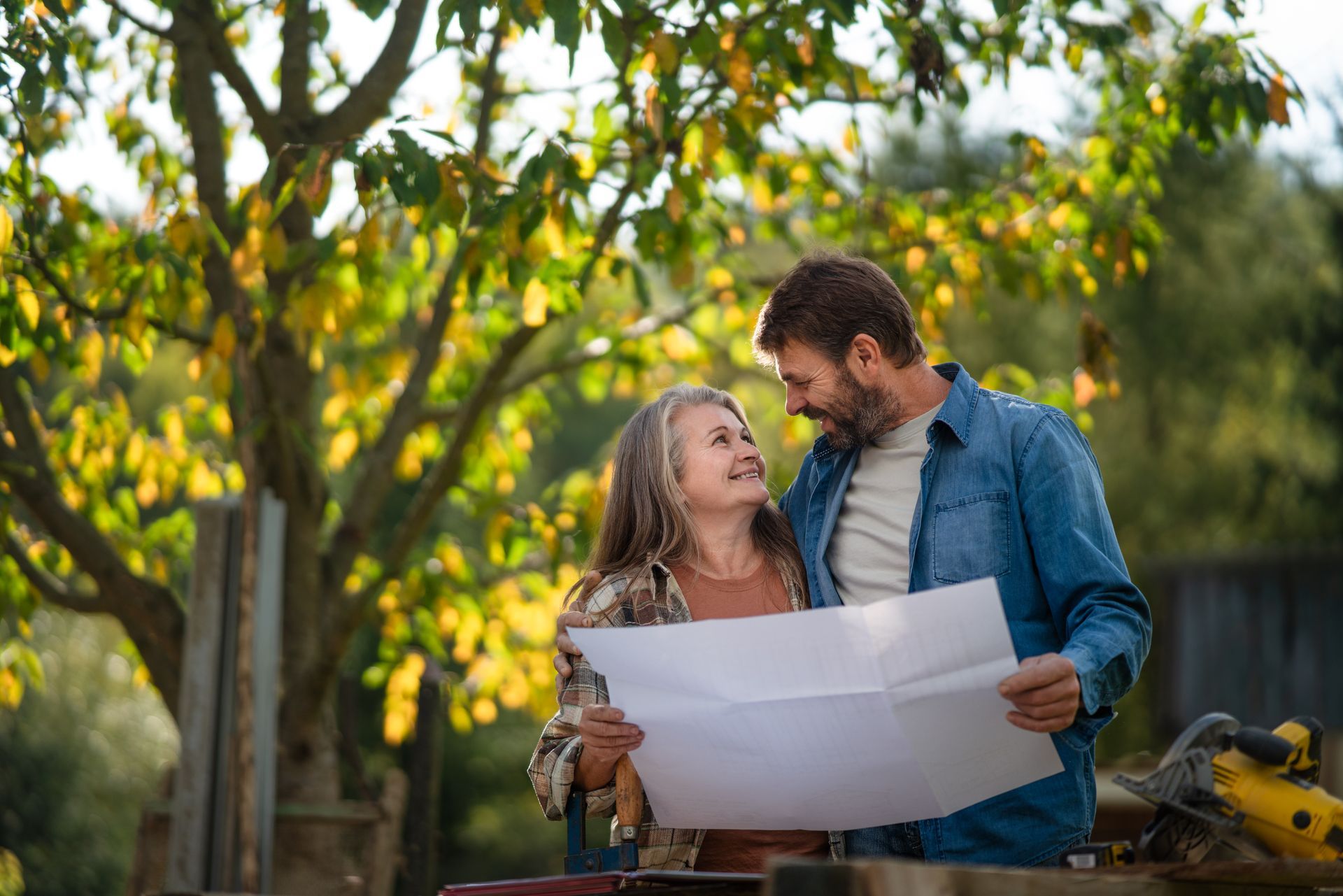 Couple looking at blueprints outdoors, smiling, with trees in the background.