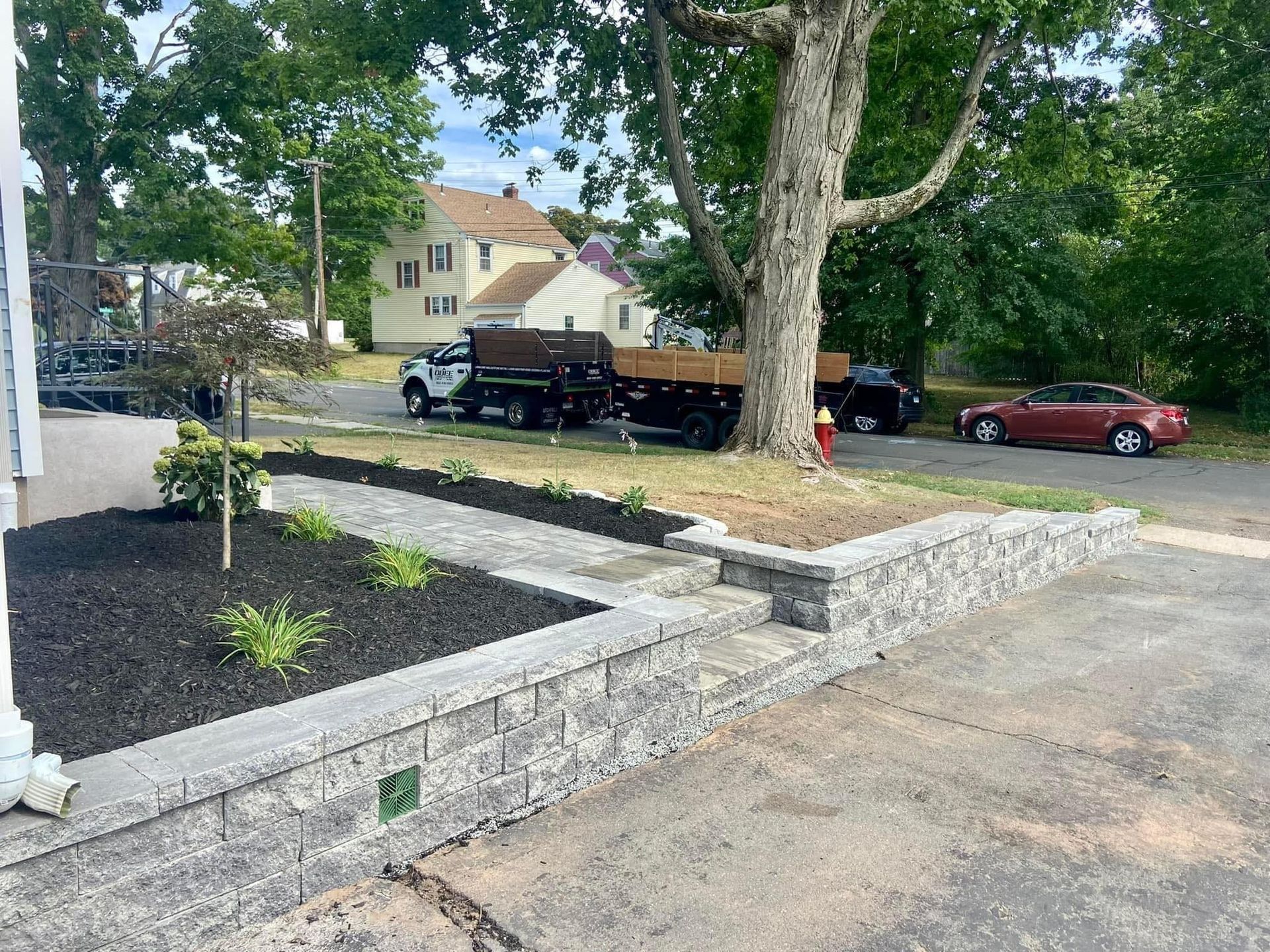 Stone retaining wall and landscaped walkway leading to a street with parked vehicles and houses in the background.