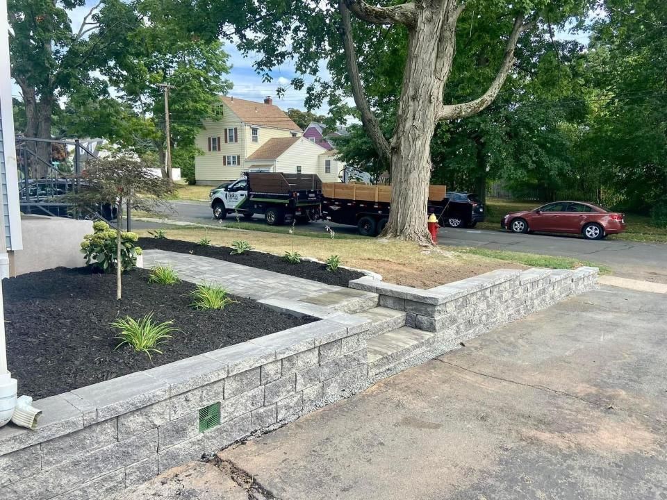 Stone retaining wall and walkway with fresh landscaping in front of a house, trucks parked nearby.