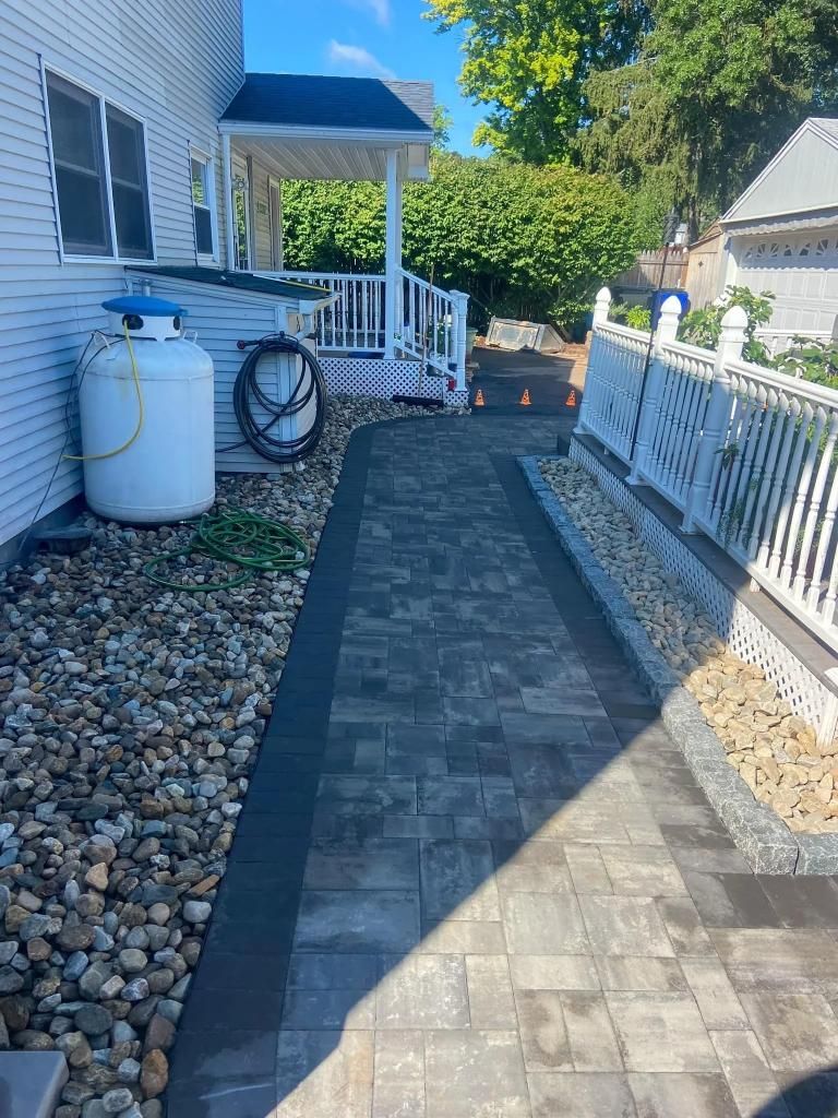 Brick pathway alongside a house, leading toward a porch with white railing. Gravel and a propane tank sit on the left.