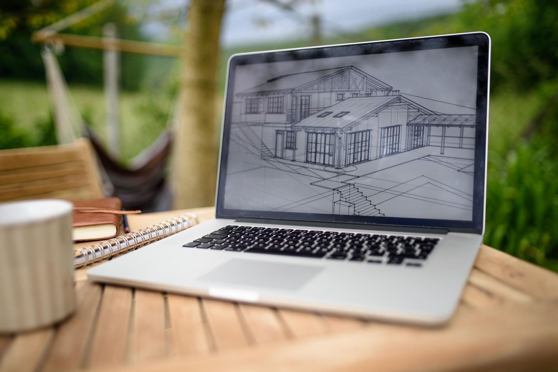 Laptop displaying house architectural drawing on wooden table outdoors.