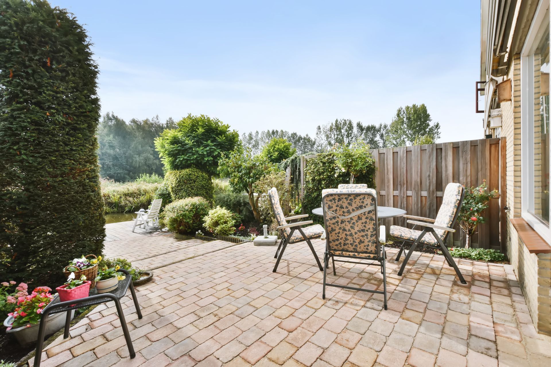 A paved patio with outdoor chairs and a small table, surrounded by garden greenery and a wooden fence on a sunny day.