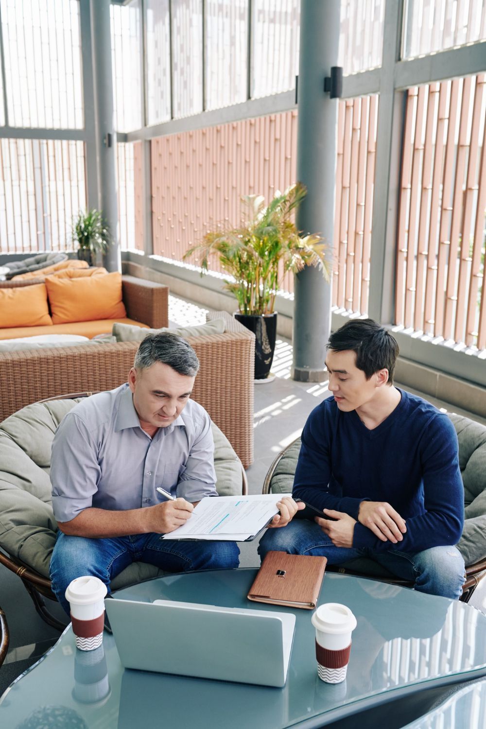 Two men review documents seated in a sunroom. One signs a paper; laptop and coffee cups are on the table.