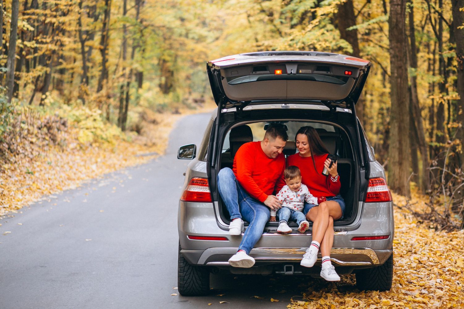 Family sitting in open car trunk on fall road.