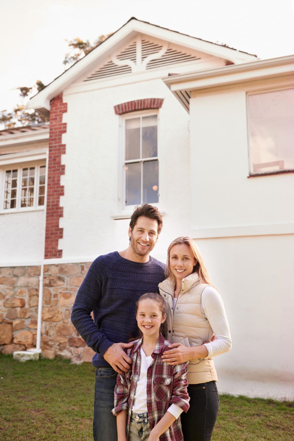 Family of three smiling in front of a white house with red brick accents.