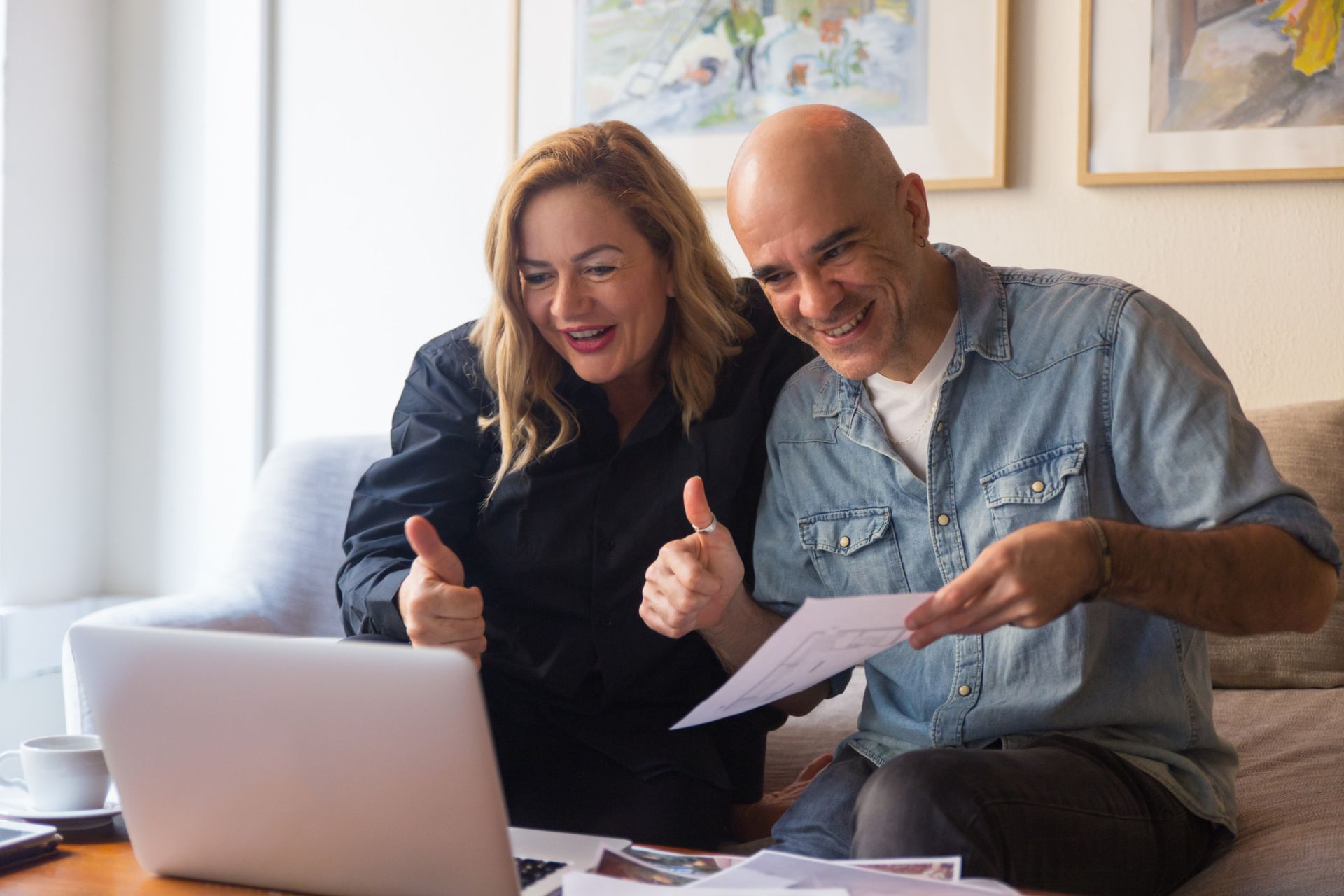 Couple on couch looking at paperwork with thumbs up and laptop.