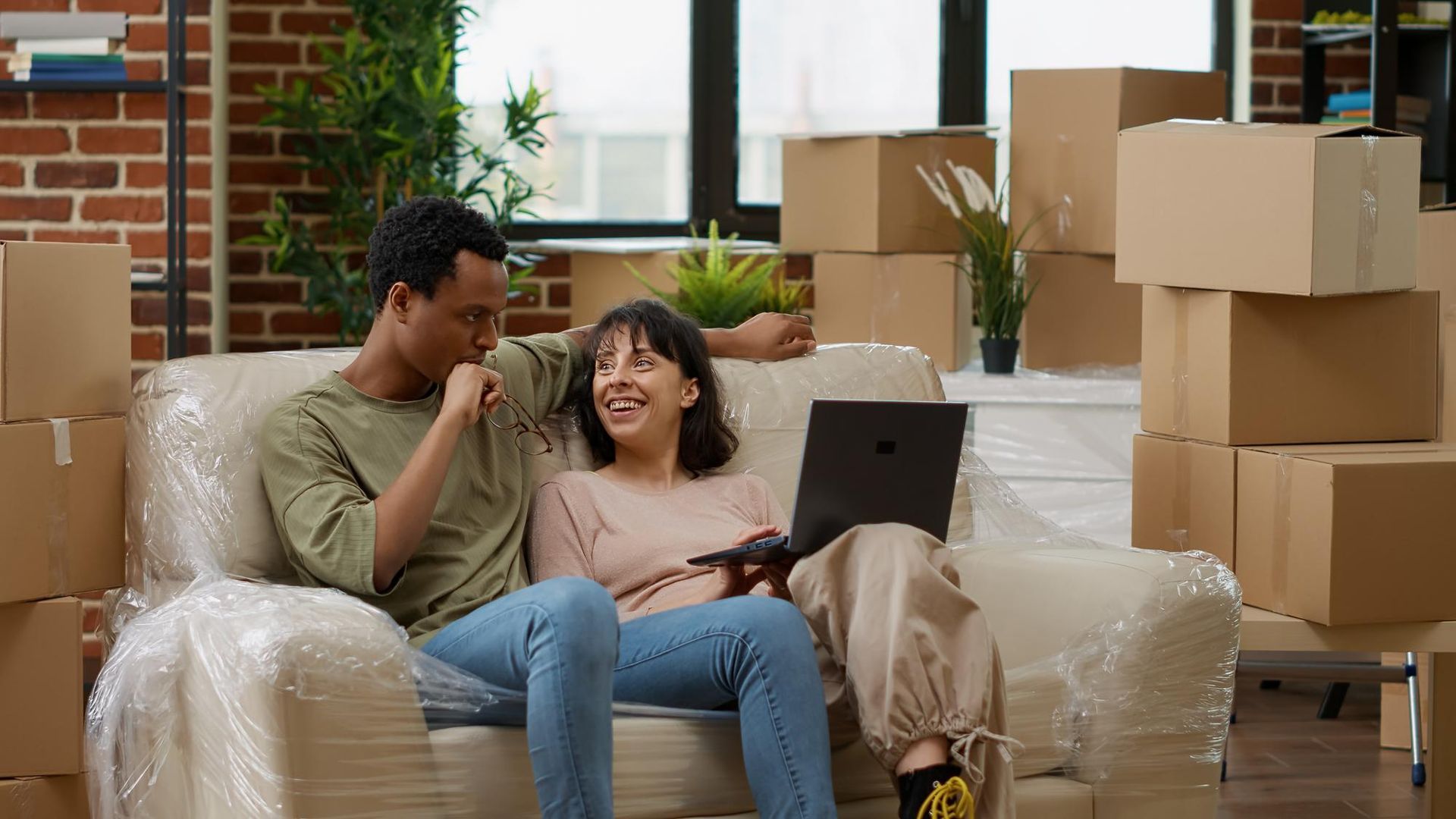 Couple on plastic-wrapped couch, looking at laptop, surrounded by moving boxes.