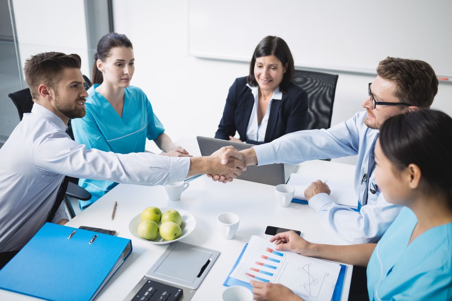 People in business attire and medical scrubs shaking hands at a table, smiling.