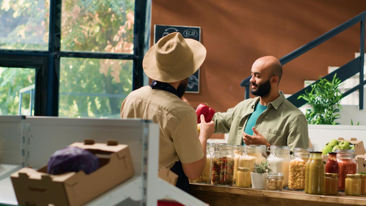 Two men in a shop, one holding out a red apple. The shop has produce and bulk food.