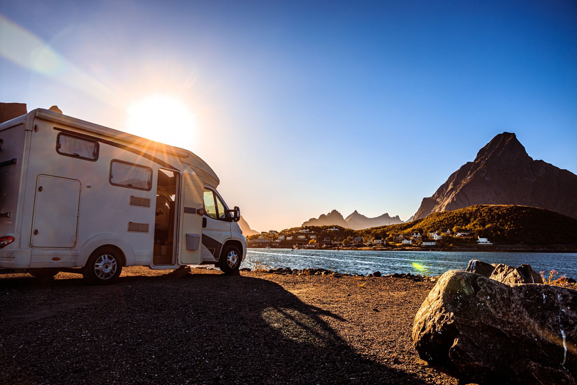 RV parked by a body of water with mountains in the background, sun shining.