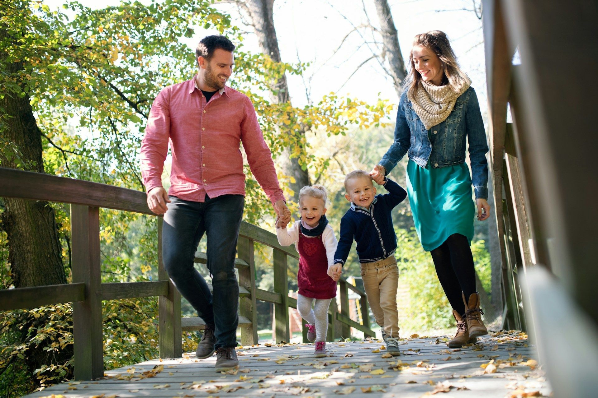 Family of four walking on a wooden bridge outdoors, holding hands; sunny day.