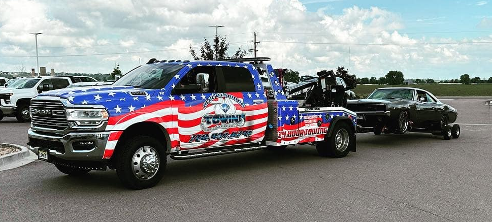 Tow truck with American flag design towing a black car on a gray asphalt surface.