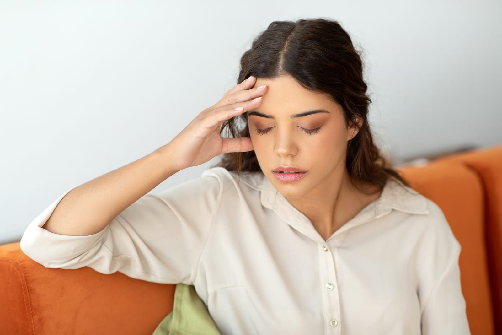 A woman suffering from a headache is sitting on a couch with her hand on her forehead.