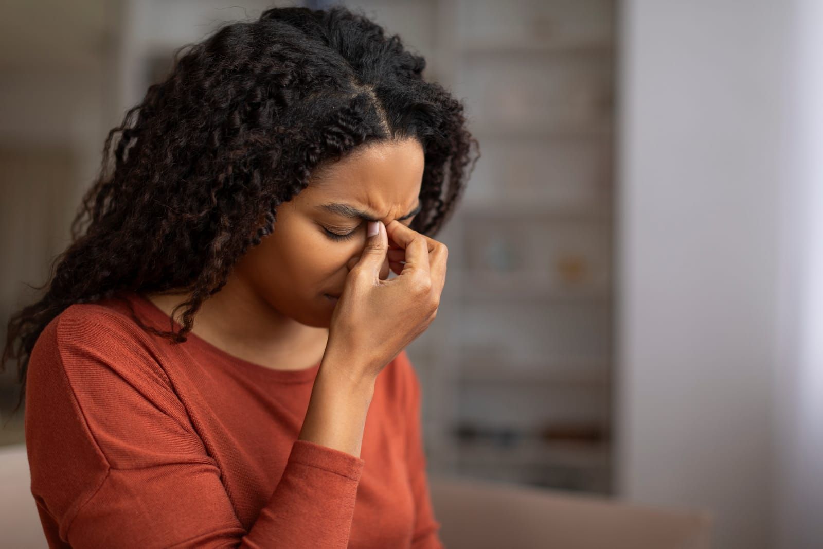 A woman is sitting on a couch holding her nose because she has a sinus headache.