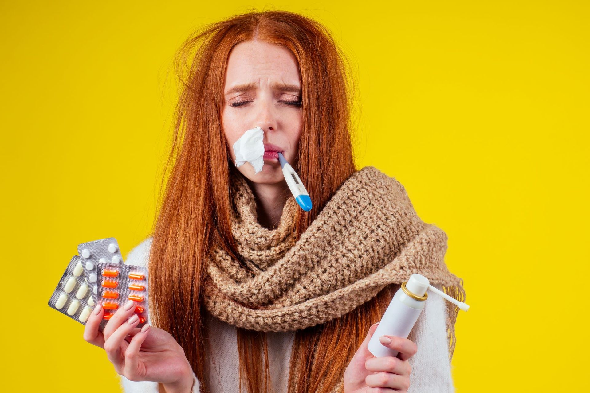 A woman with red hair is blowing her nose while holding a thermometer and pills.