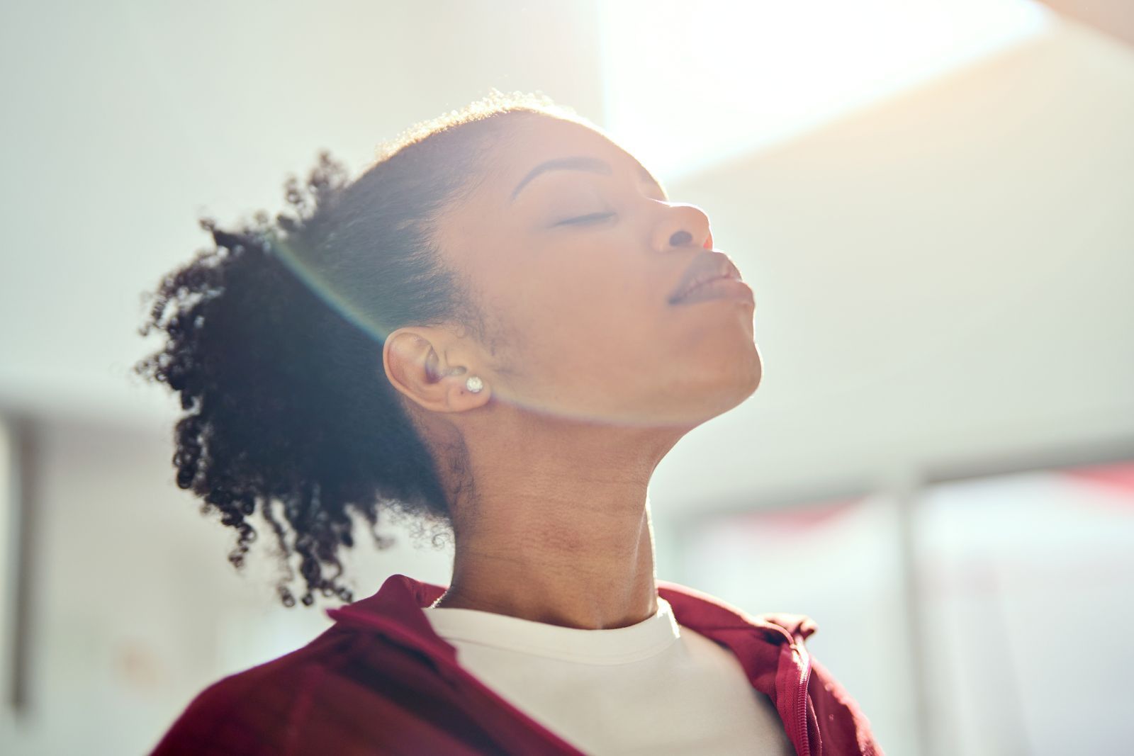 Woman with eyes closed, tilting head up toward bright light. Red jacket.
