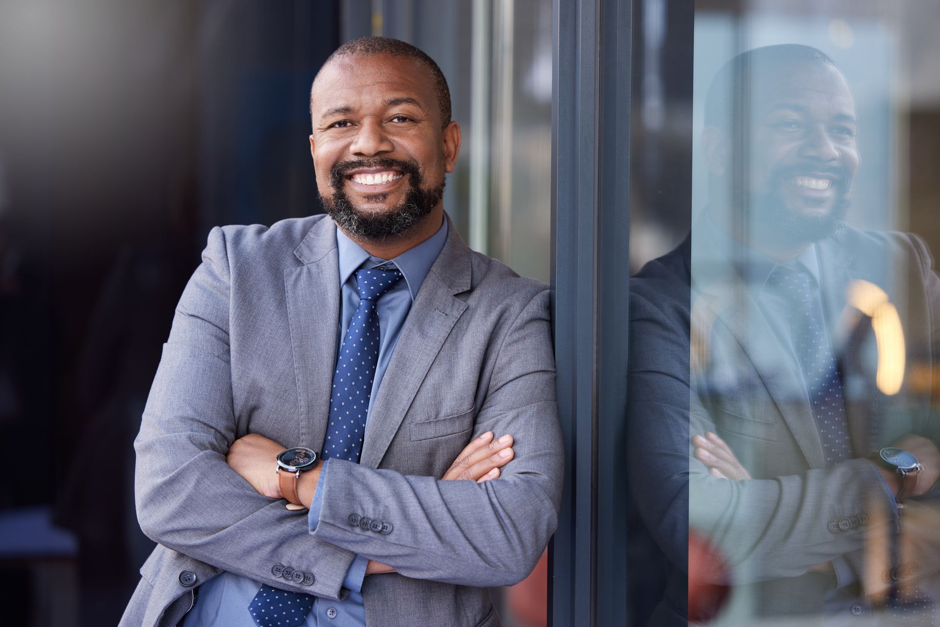 A man in a suit and tie is standing next to a window with his arms crossed and smiling.