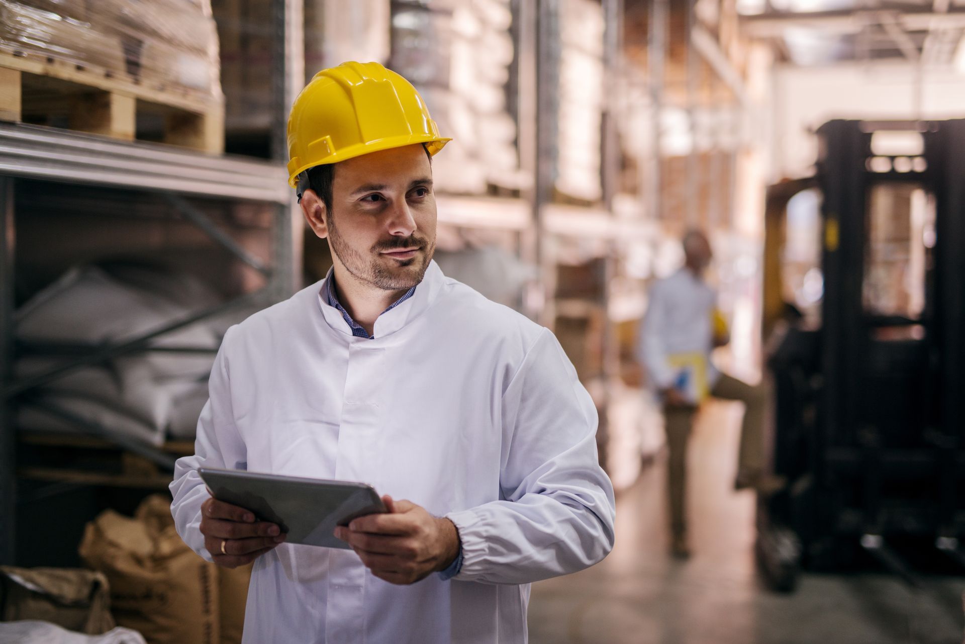 A man wearing a hard hat is holding a tablet in a warehouse.