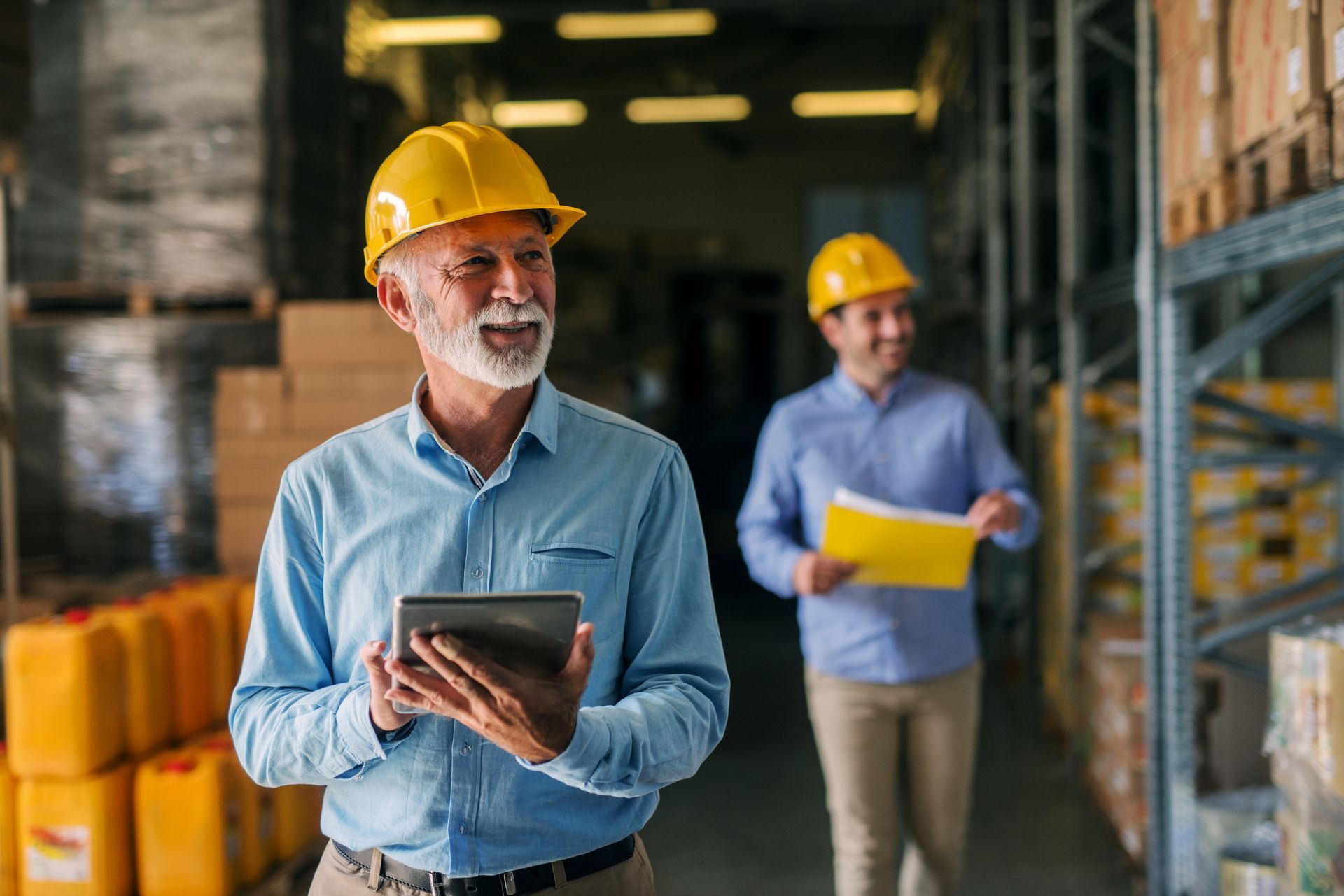 A man in a hard hat is holding a tablet in a warehouse.