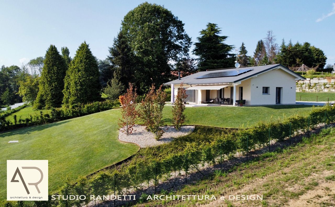 Casa moderna bianca con prato in pendenza, alberi e cielo azzurro in un soleggiato contesto rurale.