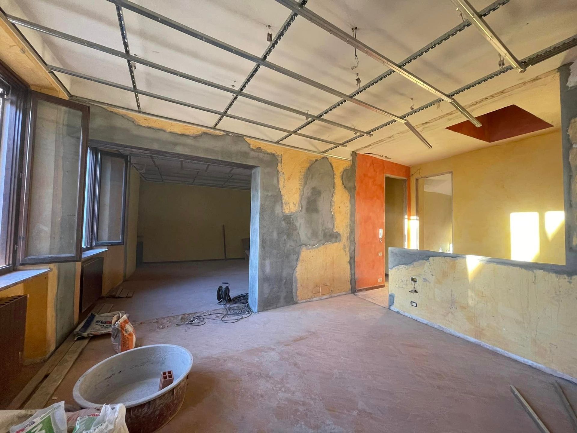 Stanza vuota in fase di ristrutturazione con soffitto a vista, pareti intonacate, finestre e materiali edili sul pavimento.