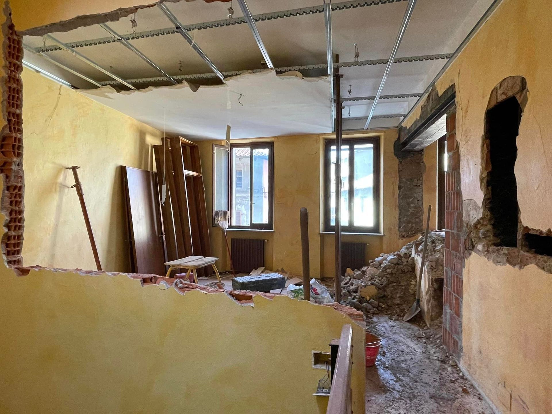 Stanza parzialmente demolita con soffitto a vista, muri rotti, detriti e finestre in pareti gialle.
