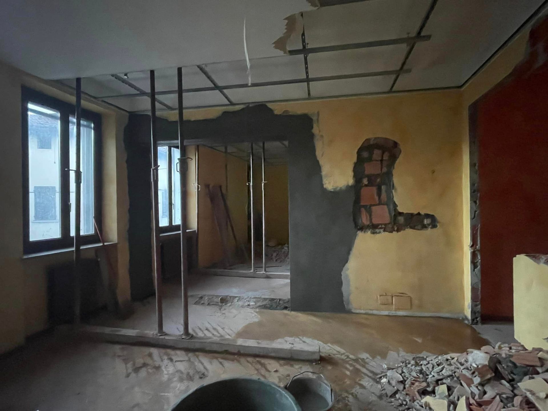 Stanza ristrutturata con struttura a vista, pareti non rifinite, finestra e detriti sul pavimento.