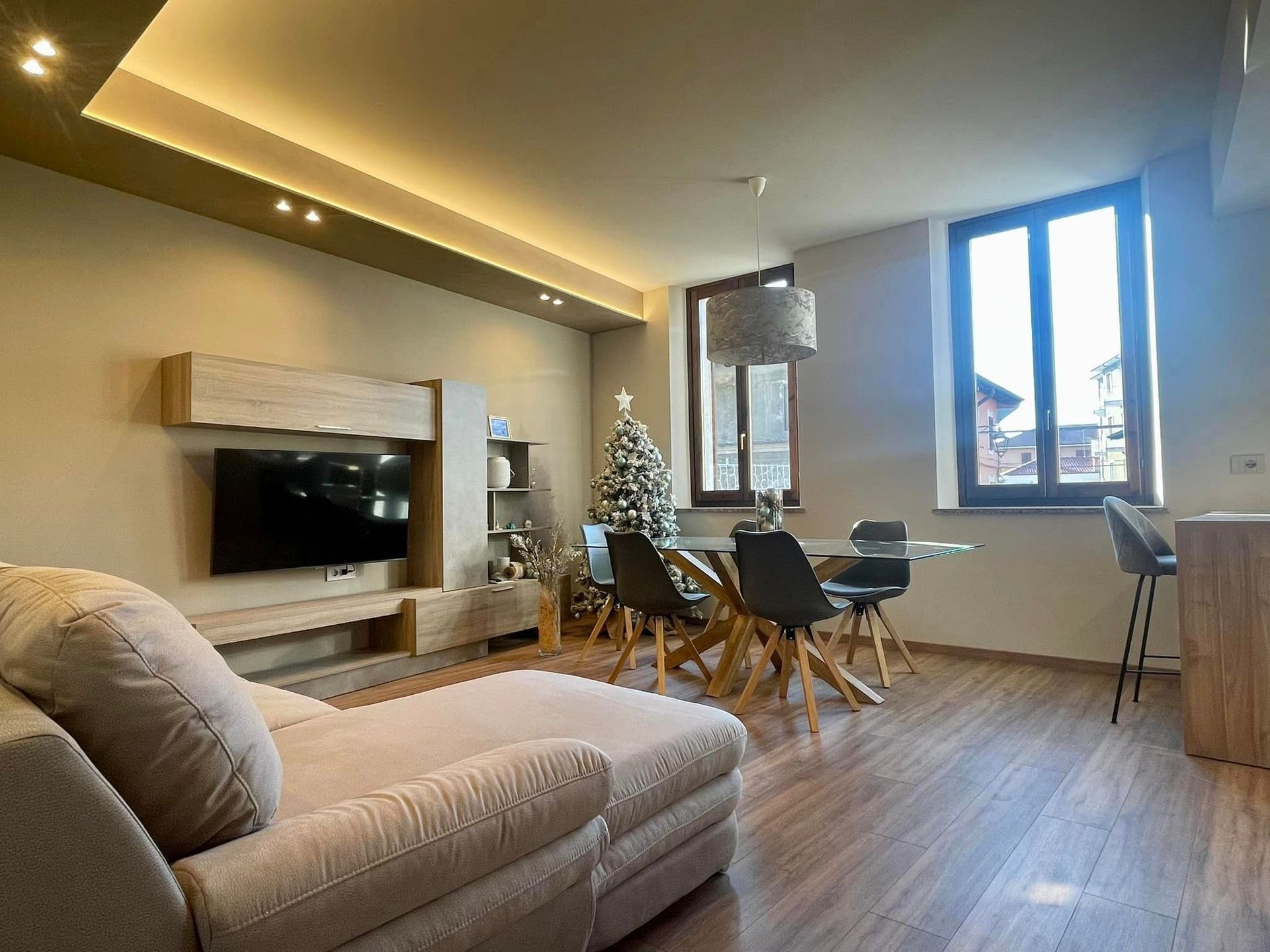 Soggiorno moderno con chaise longue beige, parete attrezzata con TV, tavolo da pranzo e ampia finestra con vista sulla città.