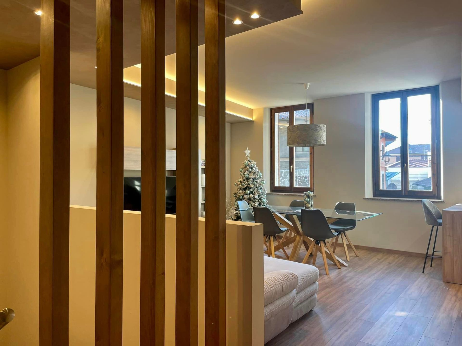 Soggiorno moderno con divisorio in legno, divano, tavolo da pranzo, albero di Natale e ampie finestre.