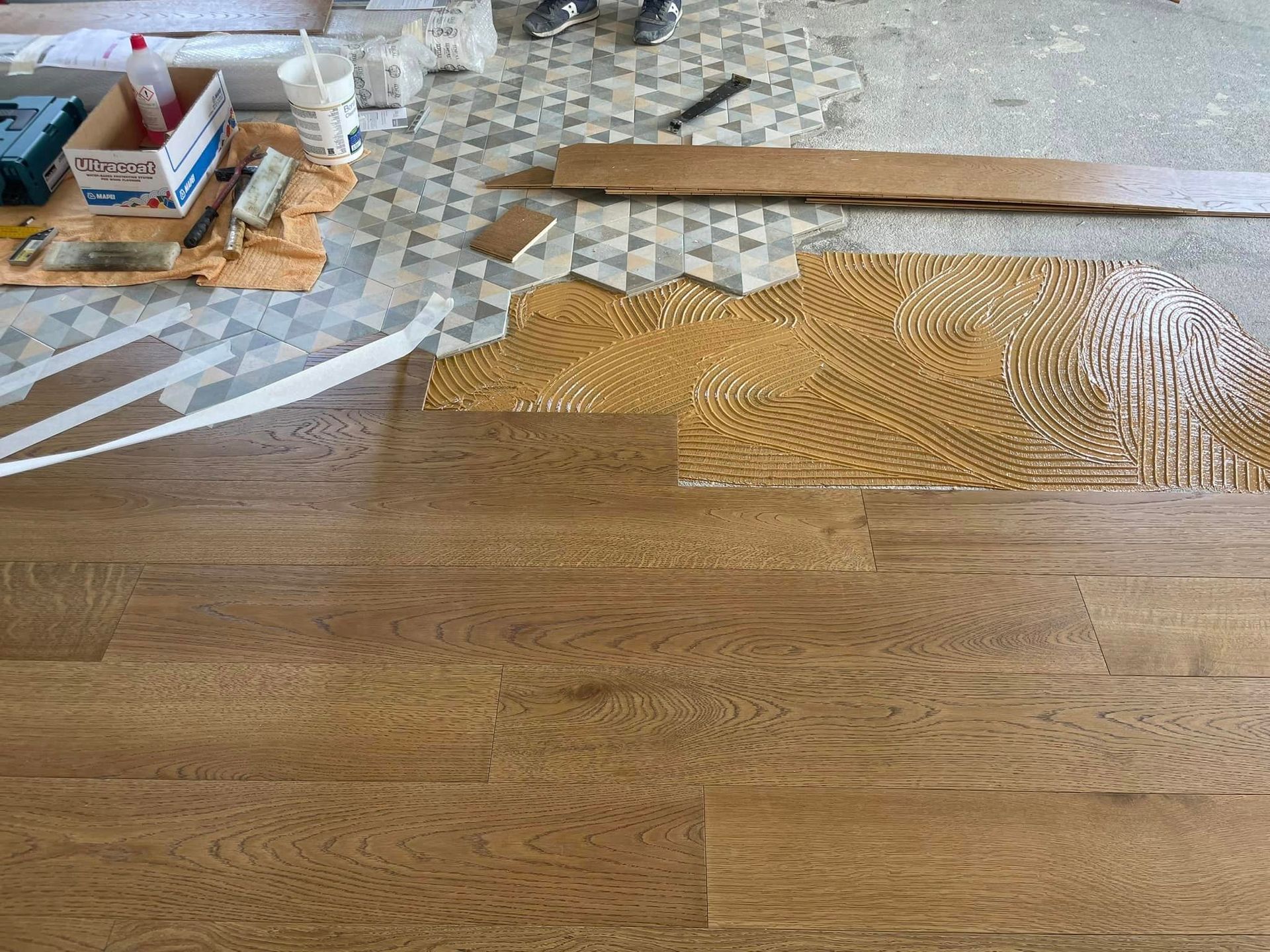 Pavimento in legno massello parzialmente installato con adesivo, attrezzi e assi di legno in una stanza in fase di ristrutturazione.