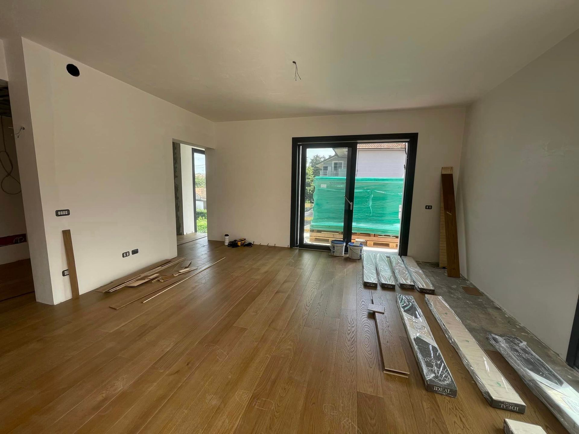 Soggiorno vuoto in fase di ristrutturazione con pavimento in legno, pareti bianche e porte finestre che danno su un balcone.