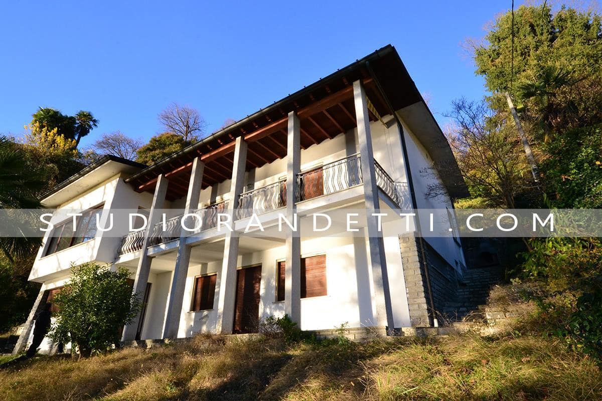 Casa bianca sulla collina con balcone in legno e tetto rosso, vista dal basso sotto un cielo azzurro.