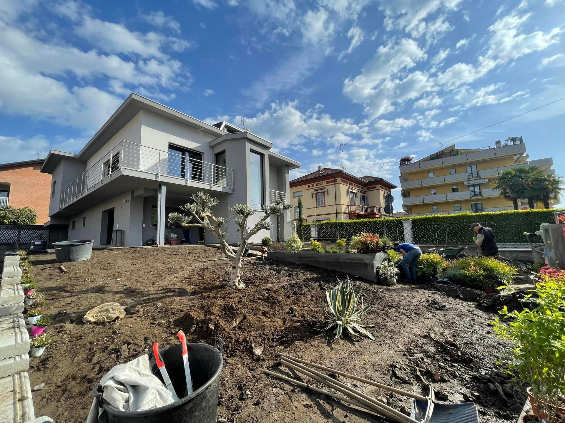 Cantiere edile nel cortile di una casa grigia, con terra scavata e operai sotto un cielo nuvoloso.