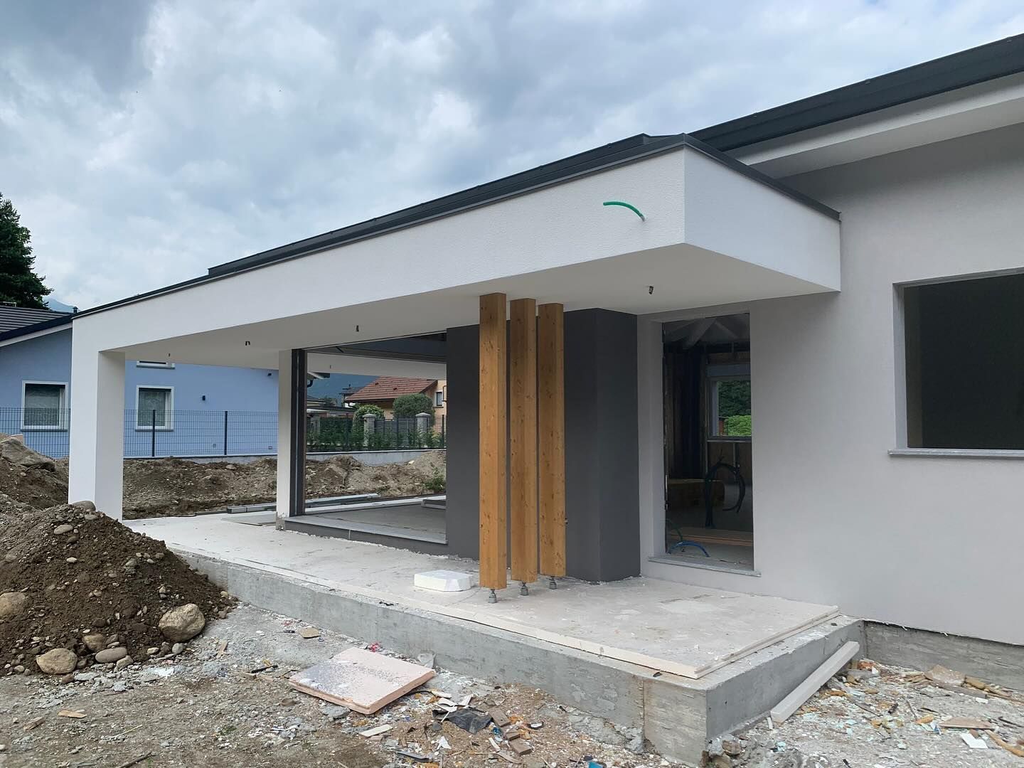 Esterno di una casa moderna con portico anteriore coperto, pareti bianche, dettagli grigi e pali di sostegno in legno in fase di costruzione.