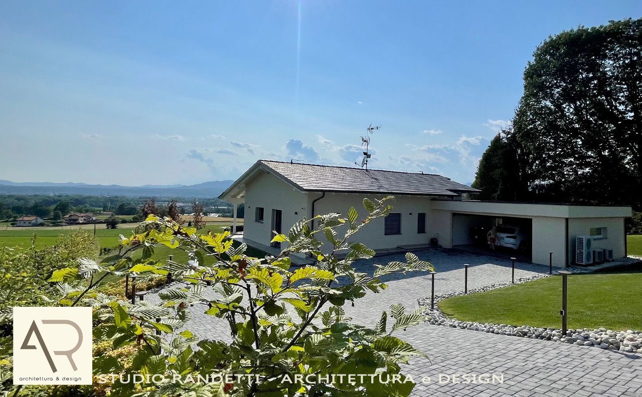 Casa moderna bianca con tetto grigio, vialetto d'accesso, garage e giardino in collina sotto un cielo azzurro limpido.
