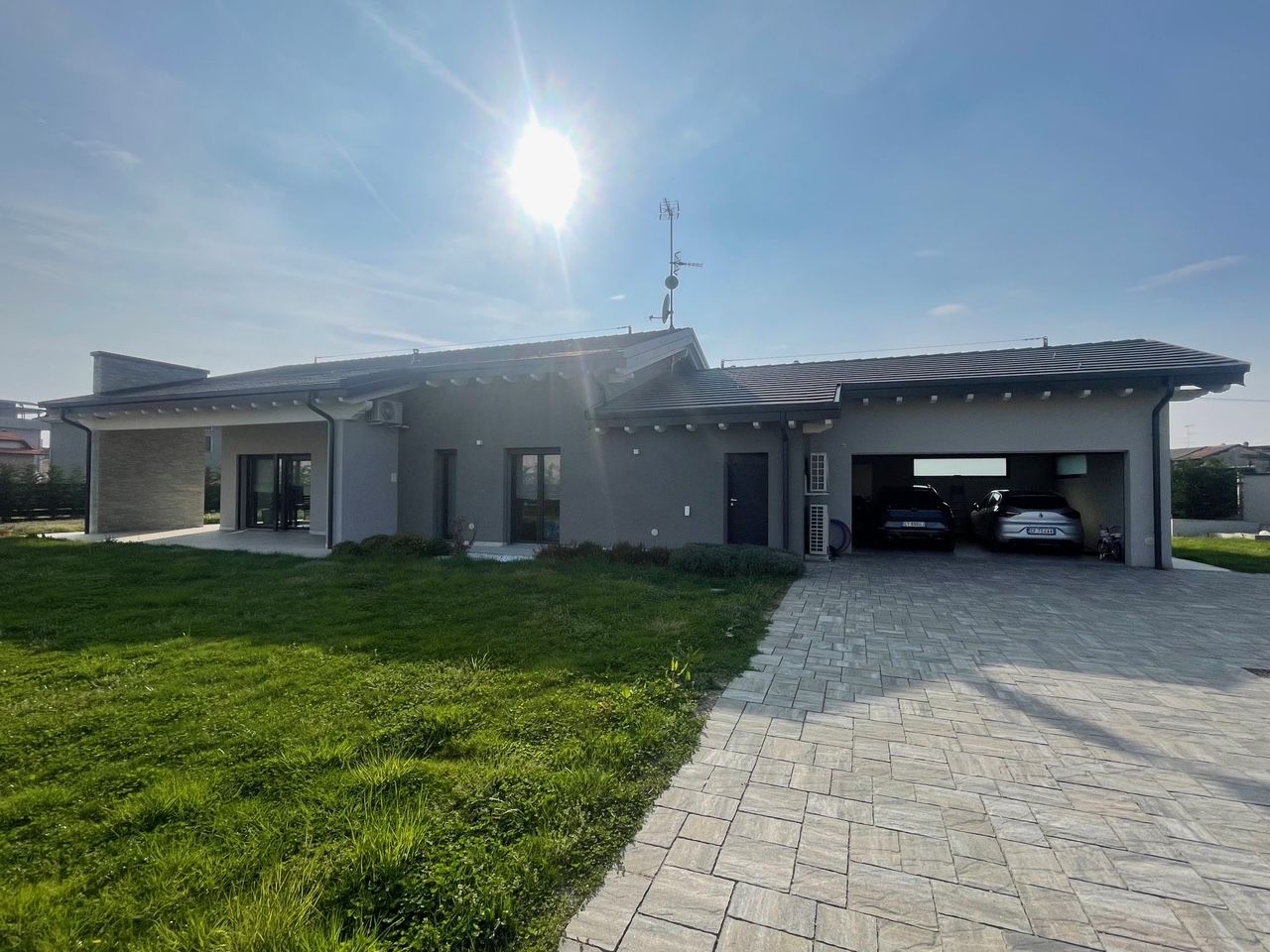 Casa grigia a un solo piano con vialetto pavimentato, prato e garage aperto, esposta a un sole splendente.
