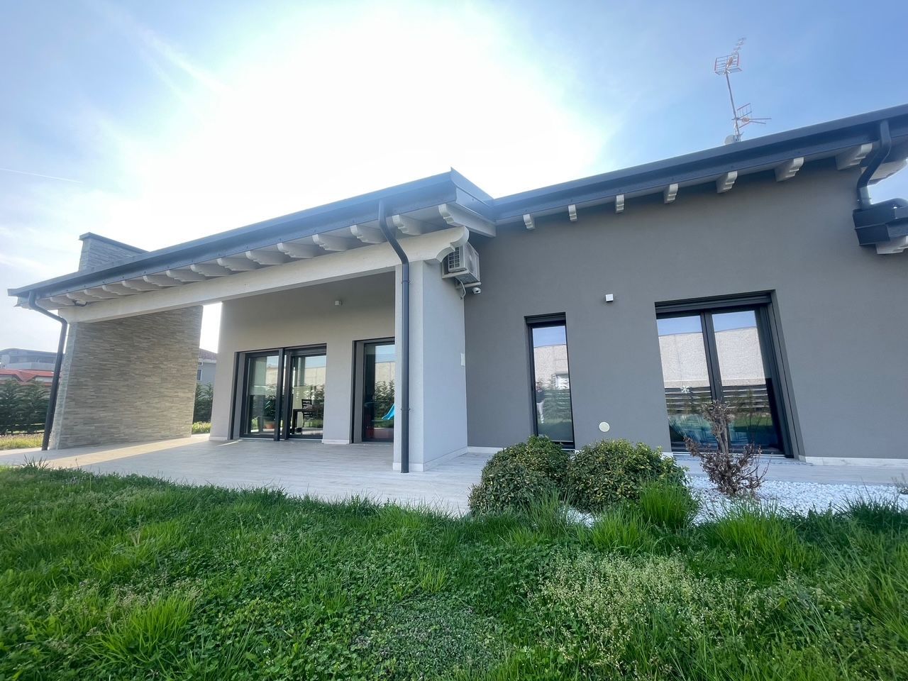 Casa moderna grigia con patio coperto, grandi finestre e prato verde sotto un cielo luminoso